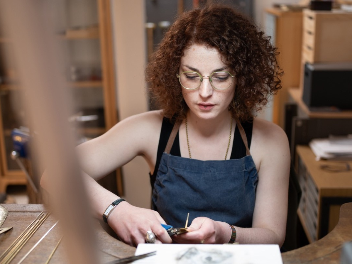 Goldbrillenträgerin mit lockigem Haar bearbeitet am Werktisch mit einer Zange ein Schmuckstück, daneben Metallstäbe und Feilen